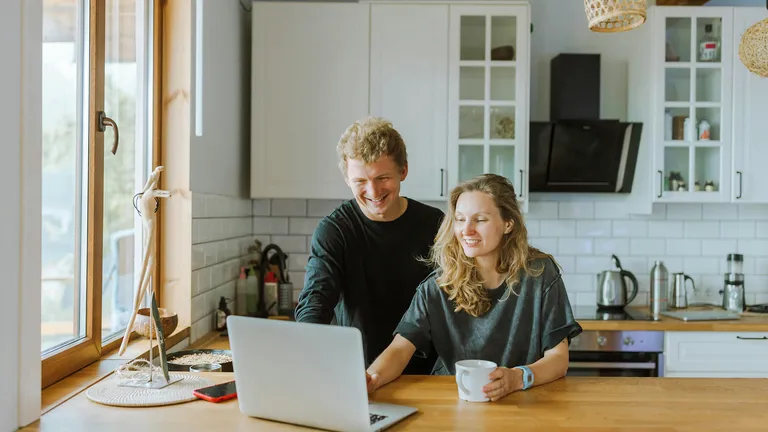 Ein junges Paar sitzt lächelnd in einer modernen Küche vor einem Laptop. Sie schauen gemeinsam auf den Bildschirm – ein Moment digitaler Alltagsorganisation oder gemeinsamer Planung.