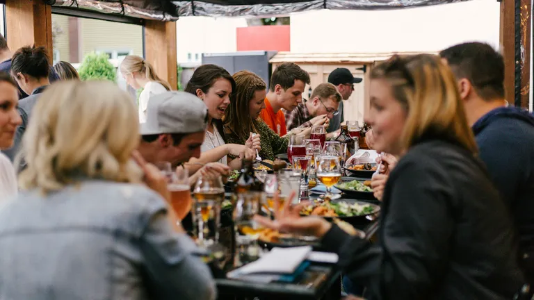 Eine grosse Gruppe junger Menschen isst und unterhält sich ausgelassen an einem langen Tisch in einem Aussenbereich – ein lebendiger Mittagstisch oder gemeinschaftliches Abendessen.